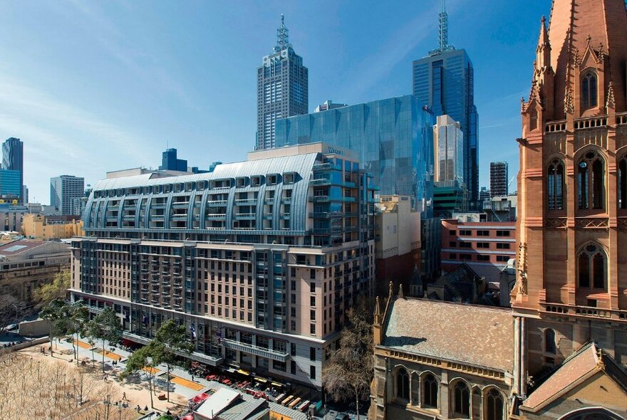 The Westin Melbourne viewed from above, showing the facade and landscaping with city skyscrapers in the background and St Paul's Cathedral next door.