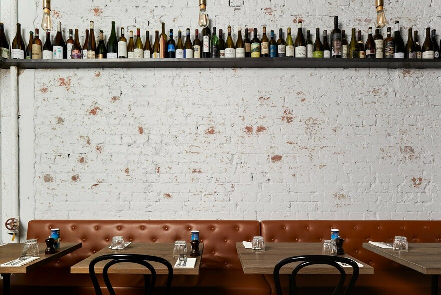Banquette seating and tables against distressed white brick wall with high shelf lined with bottles.