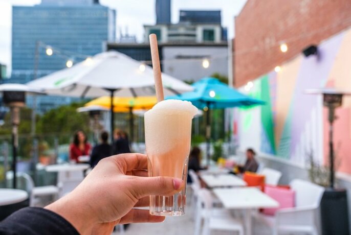Someone holding up a slushie cocktail in a colourful rooftop bar. 