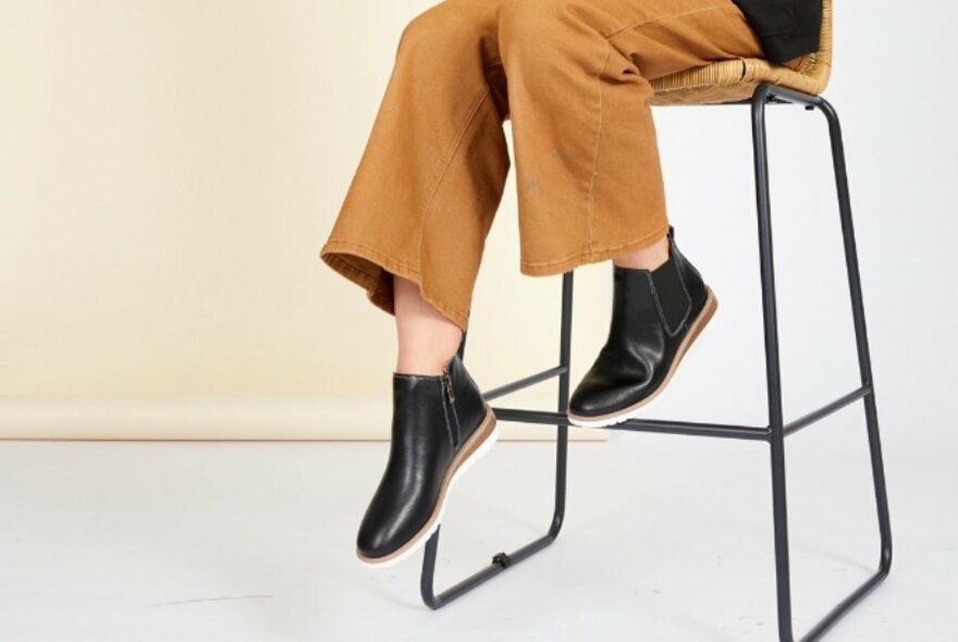 A close up of a pair of black chelsea boots with the model sitting on a high stool.