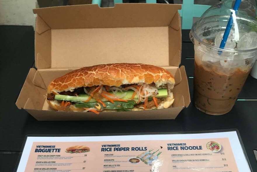 Banh mi in a cardboard takeaway box with icy drink in plastic drink container to right and menu in front.