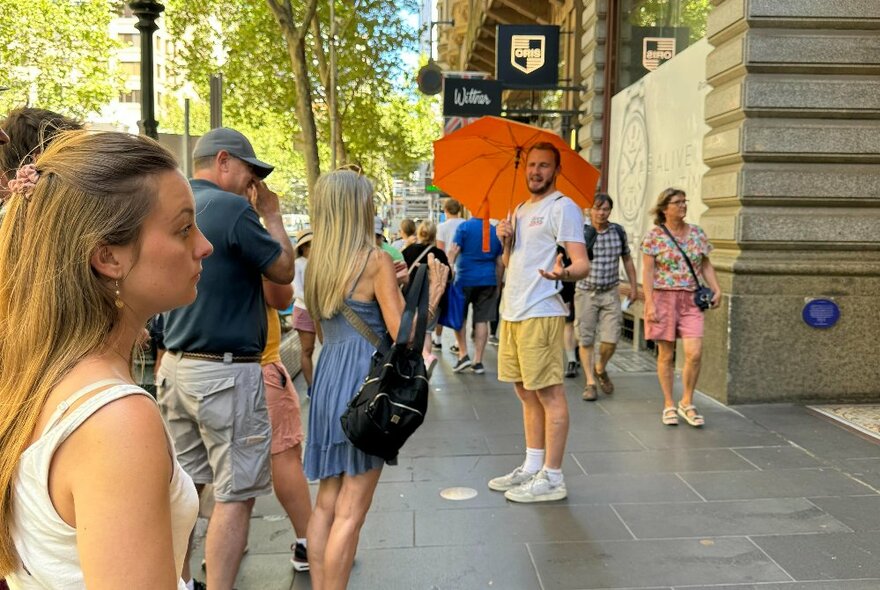 A guide holding an orange umbrella standing on a city street and talking to a small group of people on a walking tour of the city.