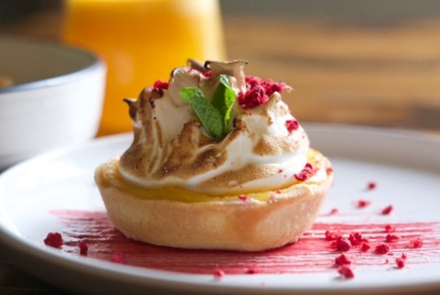 A single-serve lemon meringue pie on a white plate with a smear of freeze-dried raspberries.