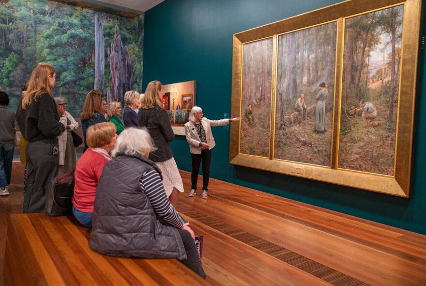 A tour guide and group in front of The Pioneer triptych by Fred McCubbin in a room with blue walls and timber floors.