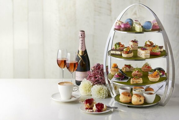 A four-tiered egg-shaped high tea stand with delectable sweet and savoury treats, alongside two glasses of wine, a wine bottle, and two tea cups. 