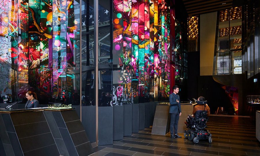 A man using a wheelchair talking to a staff member in a hotel lobby with a colourful mural.