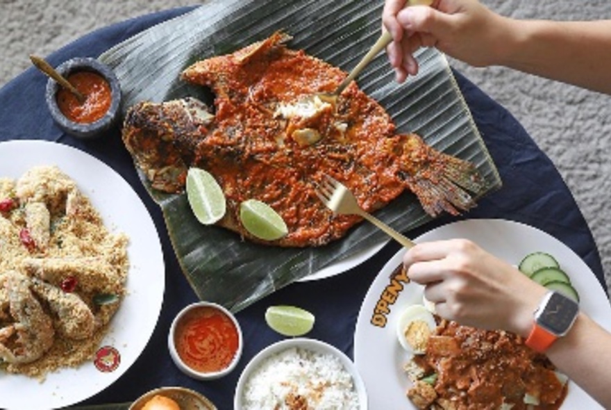 Hands using two forks to pull apart a whole baked fish on a large banana leaf, with other plates of food nearby.