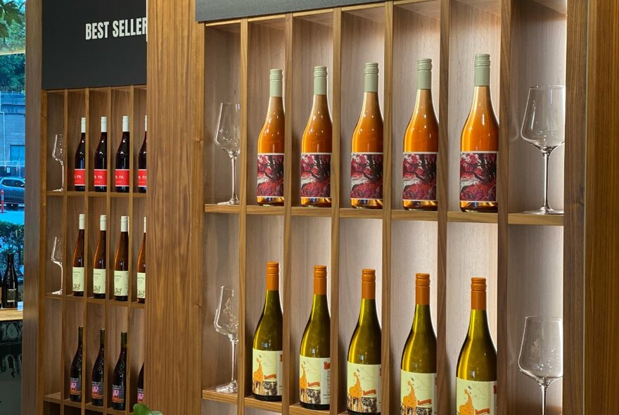 Ten bottles of white wine displayed on a wooden wine shelf with four wine glasses, and another wine shelf in the background.