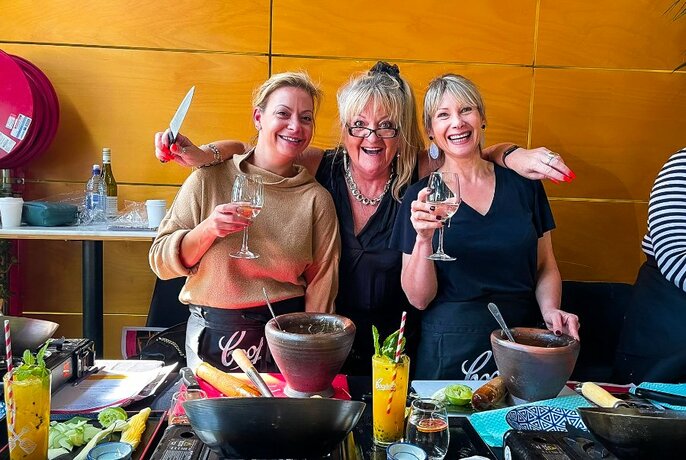 Three cooking workshop participants standing behind a food preparation table, two of them drinking a glass of wine and one holding a knife.
