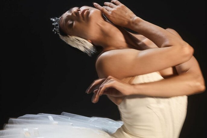 Ballet dancer in a white tutu and wearing a feathered headpiece, leaning back with arms gracefully crossed over her chest.