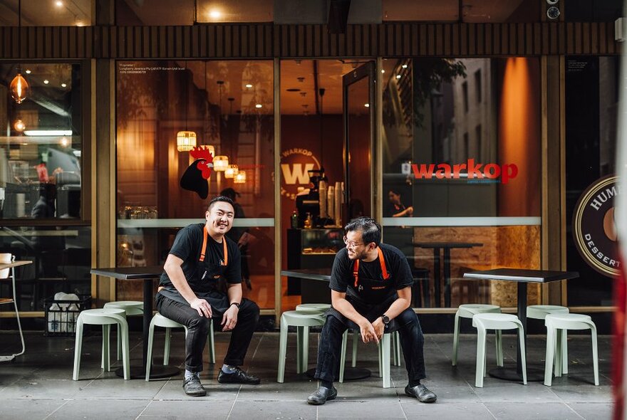 Two people in aprons relaxing on small stools outside a cafe.