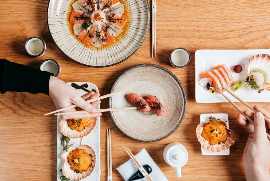 Range of Japanese dishes, viewed from above, and hands using chopsticks to select food items.