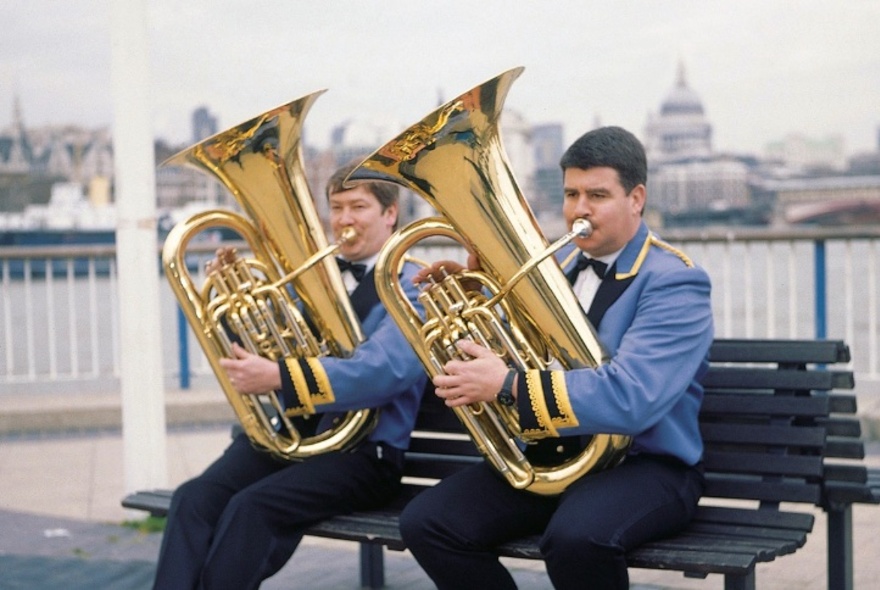 Two men in matching blue band uniforms, blowing into tubas, sitting on a bench seat outdoors, a city skyline in the background behind them.