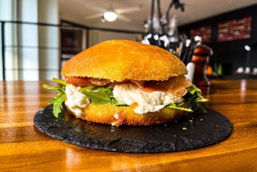 Plate on a cafe table displaying a soft bread roll filled with lettuce, soft cheese and prosciutto.