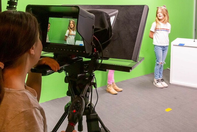 Young person looking though a film camera at two children standing against a green screen.