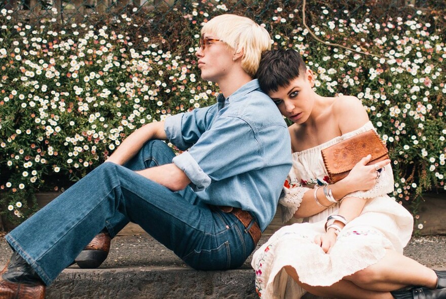Seated male model in denim with female model in an off-the-shoulder pale dress with leather bag.