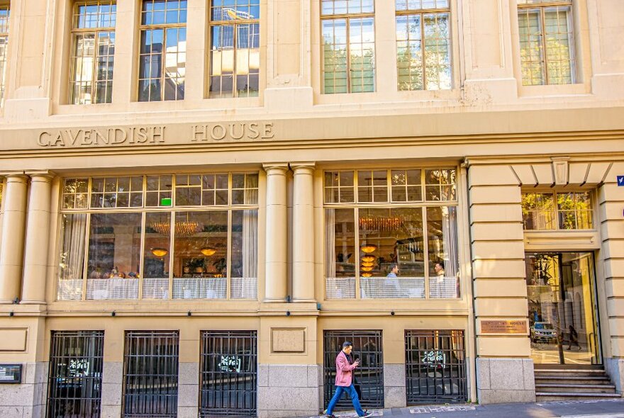 A pedestrian walking past a tall sandstone building with Cavendish House written on the side