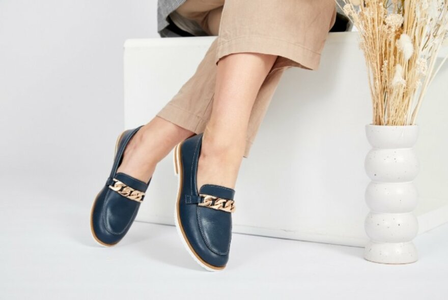 A close up of a pair of blue loafers with gold chain decoration on the end beige pants. 