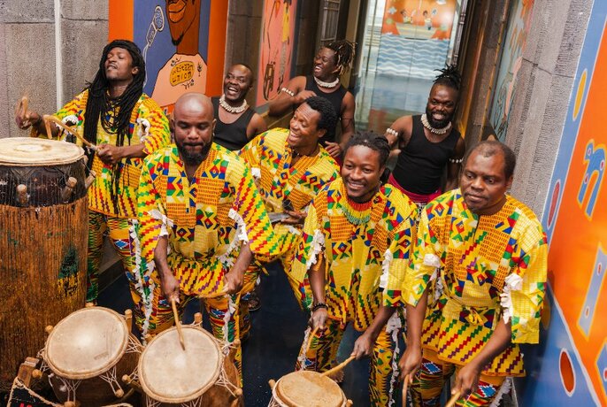 Members of the music and dance ensemble WALA, five of of them wearing colourful patterned yellow outfits, playing drums and percussion while standing.