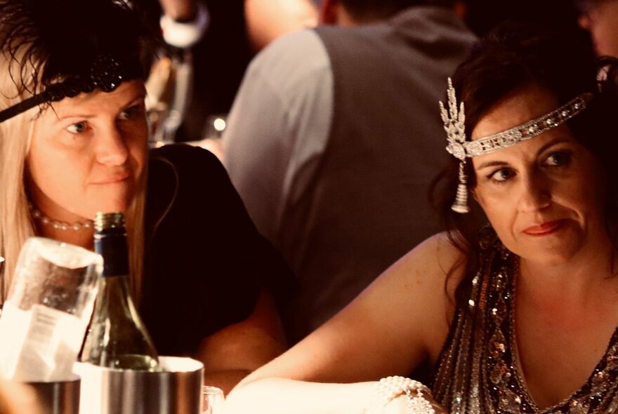 Two women seated at a table with wine, wearing 1920s-inspired jeweled headpieces and earrings.