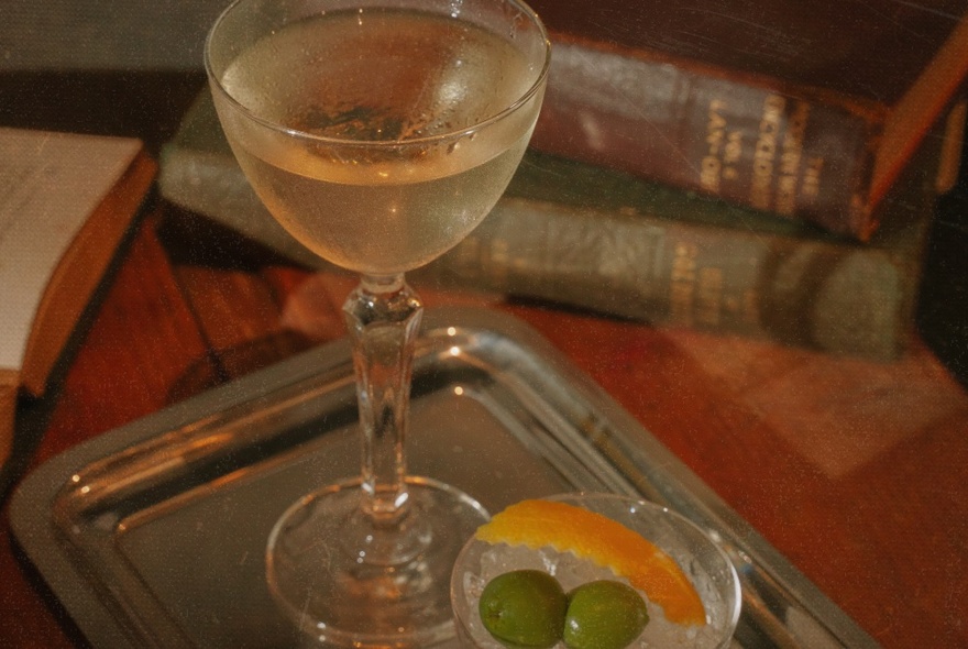 A martini cocktail, with a small bowl of olives next to it, vintage books in a pile behind it.