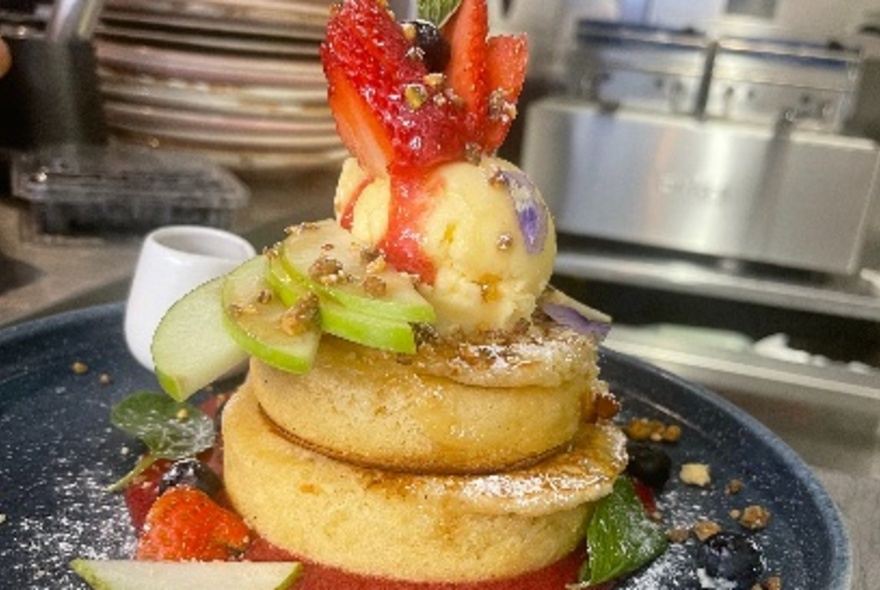 Stack of fluffy pancakes topped with a scoop of ice-cream, strawberries and slices of apple, served on a dark plate.