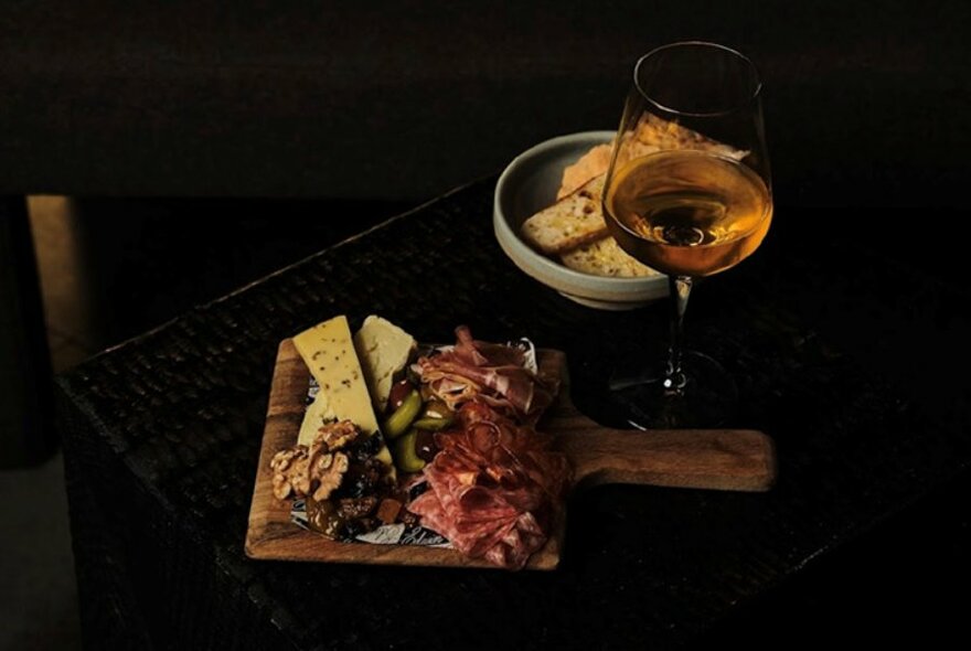A small wooden board with an antipasto selection, a glass of wine and a bowl of crackers on a table.