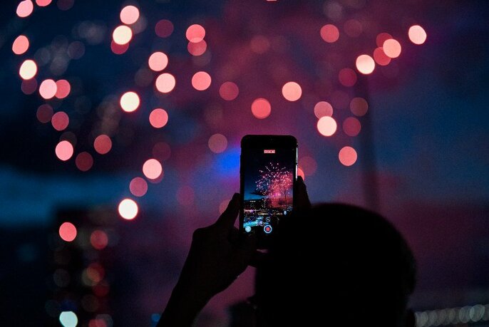 Person holding a smart phone and taking a photo of the night sky with fireworks.
