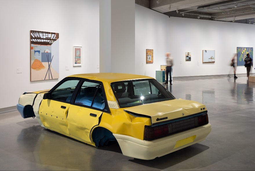 Interior of a large art gallery showing artworks on the walls, and a large yellow car sculpture in the foreground.