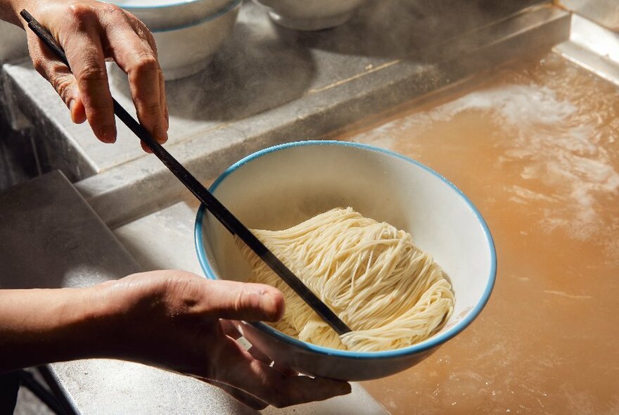 A person adding some freshly cooked noodles to a bowl with a chopstick, the boiling water in the background. 