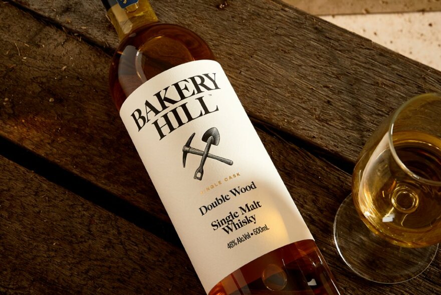 A bottle of Bakery Hill whisky lying flat on a wooden table next to a glass.
