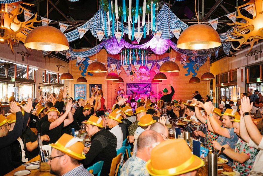 A large crowd of people wearing Oktoberfest hats and costumes, seated at long tables inside a festive looking beer hall, enjoying food and drinks.