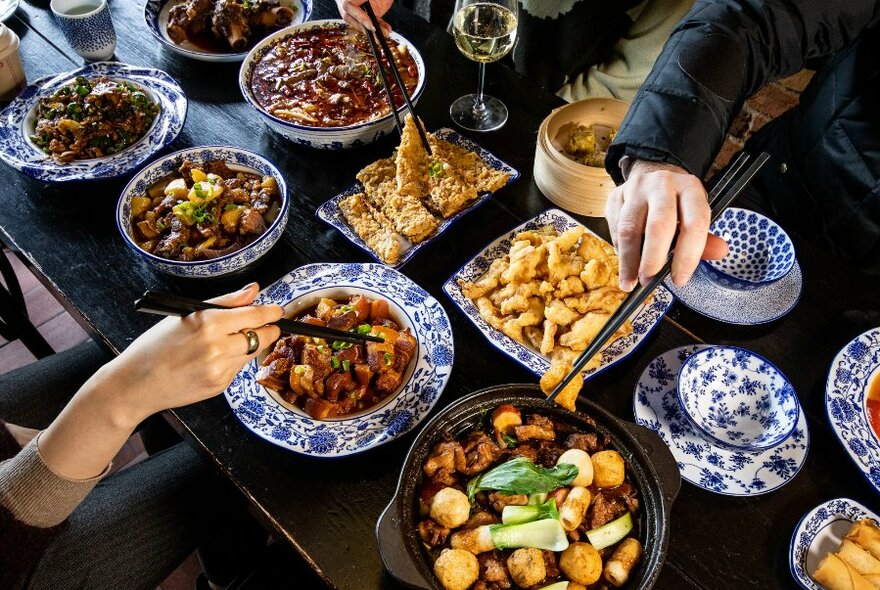 Black table laden with dishes; several hands with chopsticks over the food.