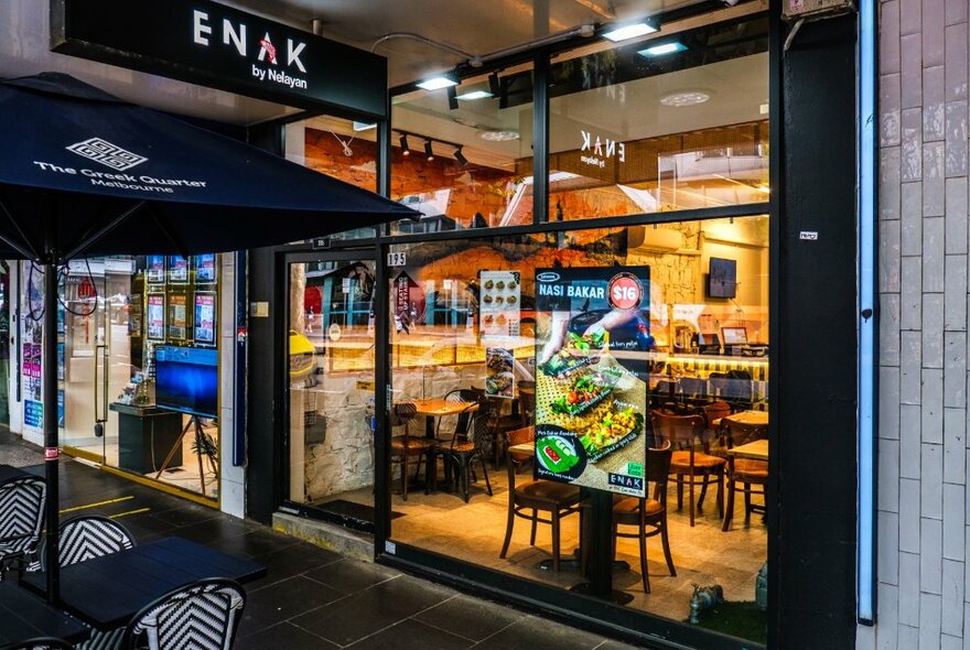 The glass exterior of Enak restaurant showing outdoor seating on the footpath, shop signage and a poster on the window.