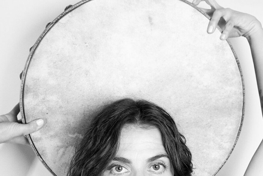 The top of a woman's head showing just forehead, eyes and hair, holding a large tambourine behind her head; black and white image.