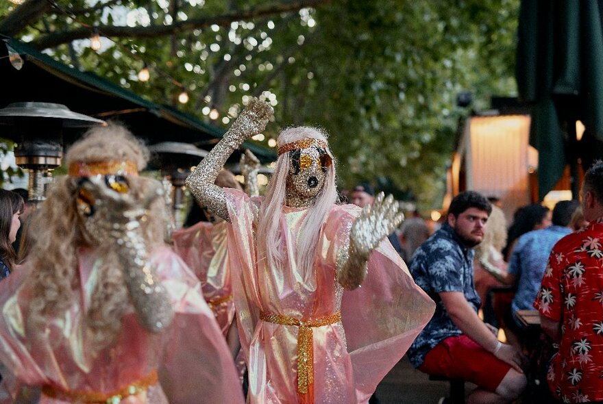 Performers at an outdoor bar dressed in glittery makeup and pink flowing gowns with capes.