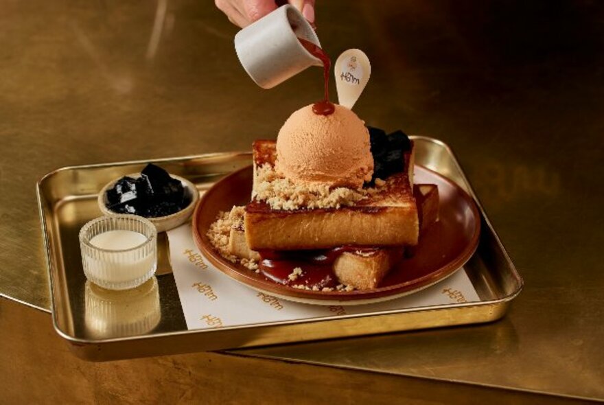 A tray resting on a table with a plate of dessert and someone pouring a sauce over it. 