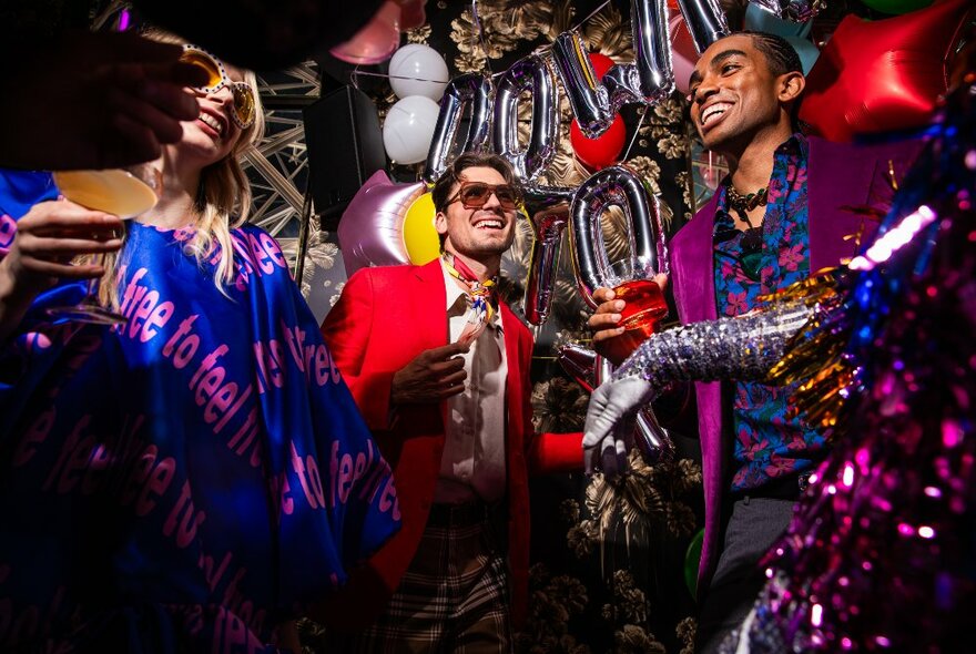 A few people dressed up, smiling, dancing and  having a fun time drinking cocktails, with sparkling decorations and balloons around them.