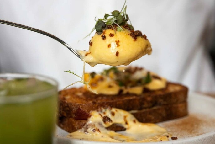 Poached egg on a spoon held above toast with a green juice to the left.