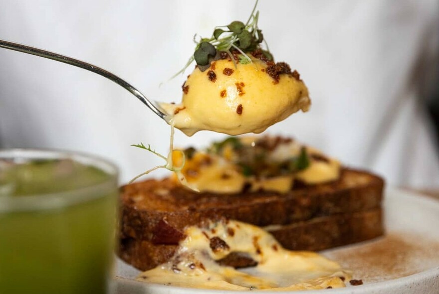 Poached egg on a spoon held above toast with a green juice to the left.