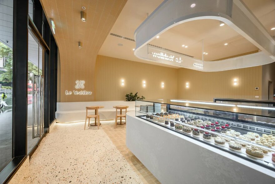 Sleek, modern interior of a cake shop with terrazzo floors, a large glass display counter and some small stools and table against the wall.