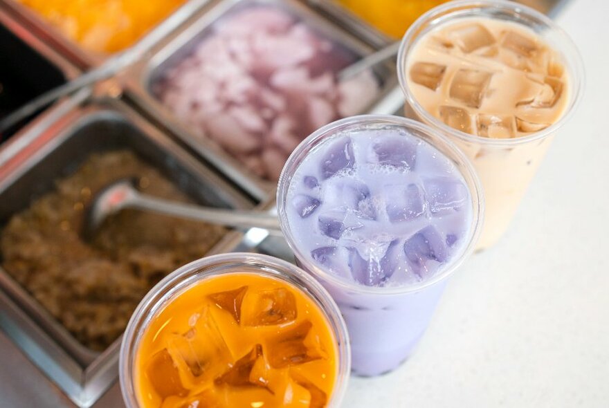 A selection of colourful boba drinks and various toppings in the background. 