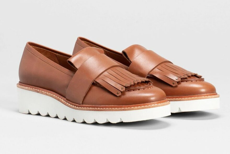 A pair of brown loafers with a fringe detail on the front and puffy white soles.