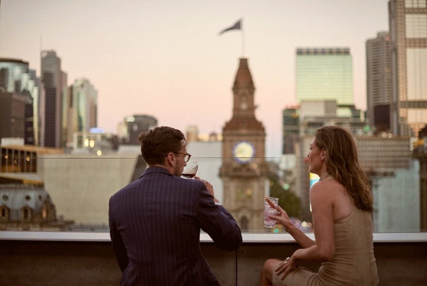Two people seated at a rooftop bar with a view of Melbourne's GPO in the background.