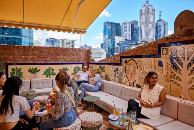 People seated at a rooftop bar under a yellow awning with the Melbourne skyline in the background. 