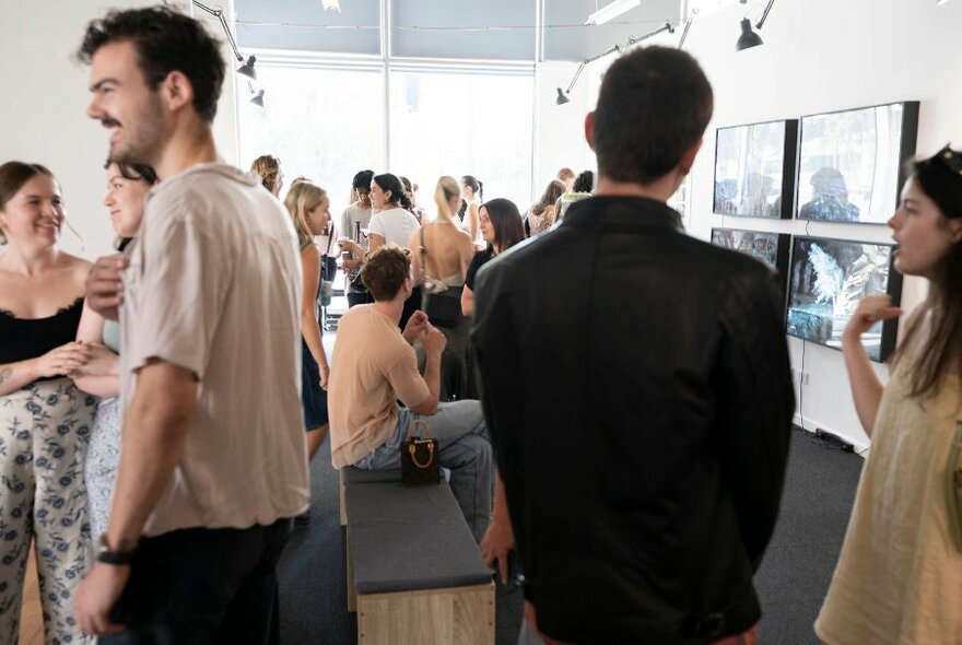 A gallery space filled with people mingling and viewing an exhibition displayed on the walls. 