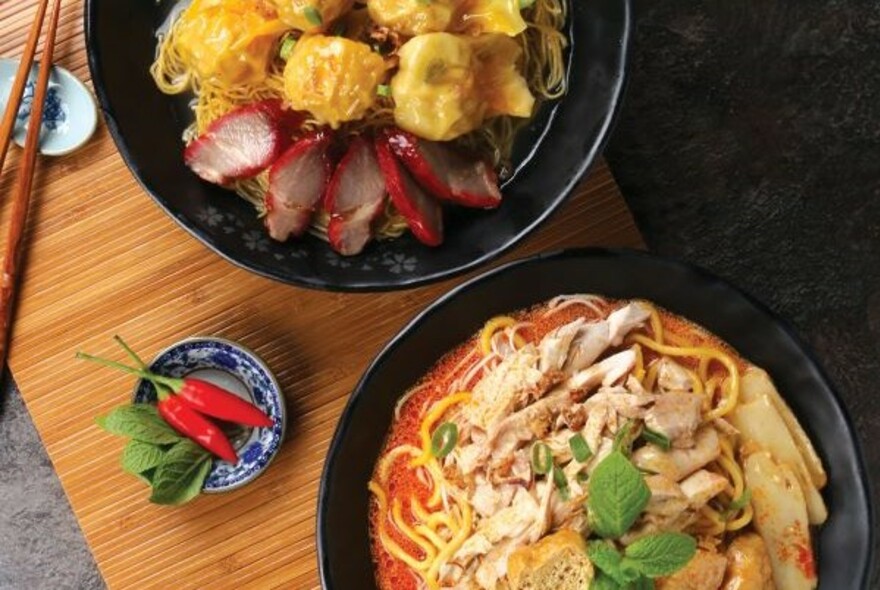 Wooden table with black bowls of meat and noodle dishes, with red chillies in a blue bowl.