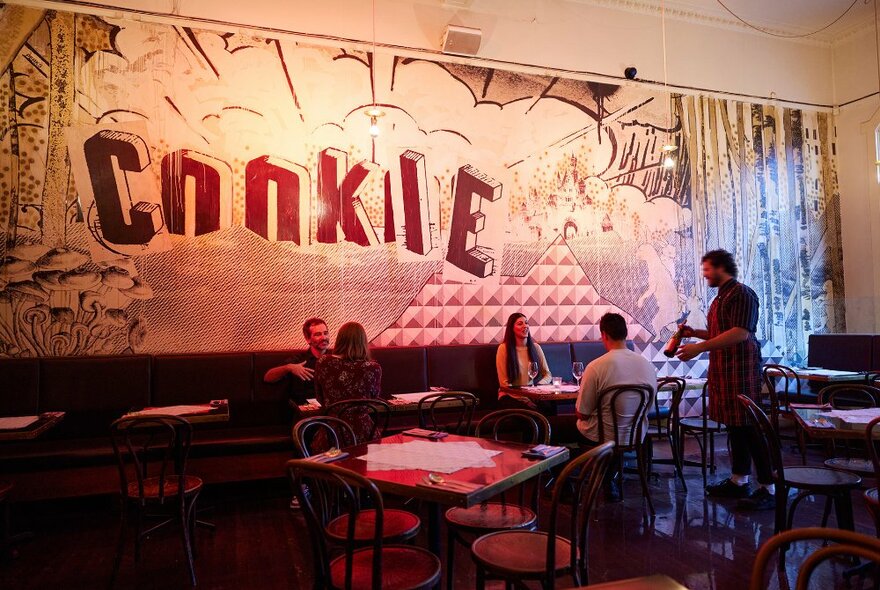 A waiter showing a bottle of wine to a couple in a dim restaurant with a mural on the wall that says Cookie.