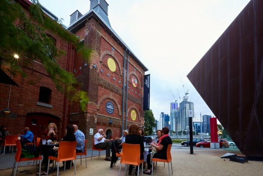People seated at cafe tables outside old red brick building housing Malthouse Theatre.