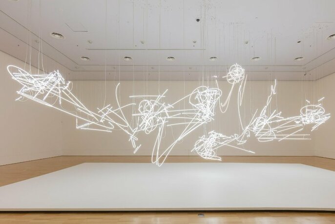 Installation view of a suspended neon sculptural piece made up of curved and squiggly white neon lines, hanging from a ceiling as if floating in a large airy space.